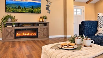 Una sala de estar acogedora con chimenea, un televisor montado en la pared y un espacio de descanso cómodo con un sillón azul y cojines.