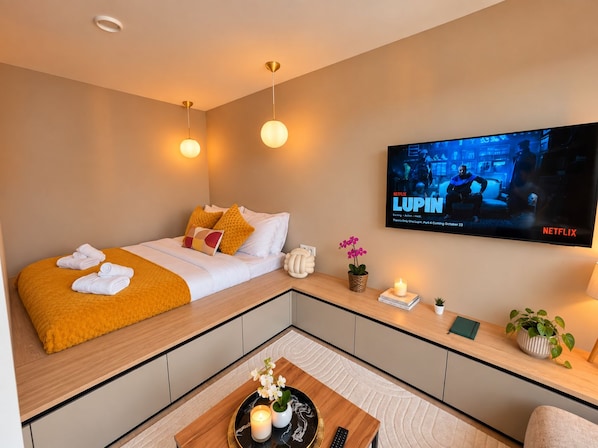 Quarto de hotel moderno com cama, TV exibindo um programa da Netflix, mesa de cabeceira com planta e um pequeno espaço de estar com mesa e cadeiras.