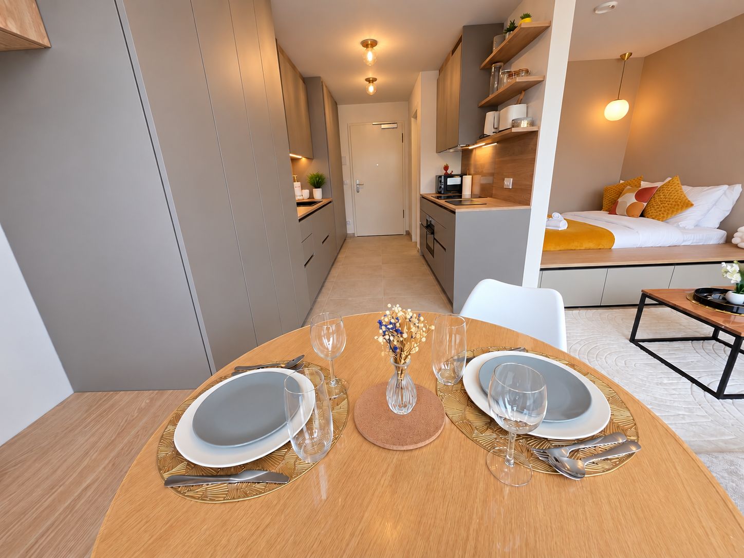 A dining area with a table set for two, a kitchen in the background, and a living area with a sofa and cushions.