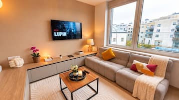 A modern living room with a flat-screen TV, a sofa, a coffee table, and a view of the city through large windows.