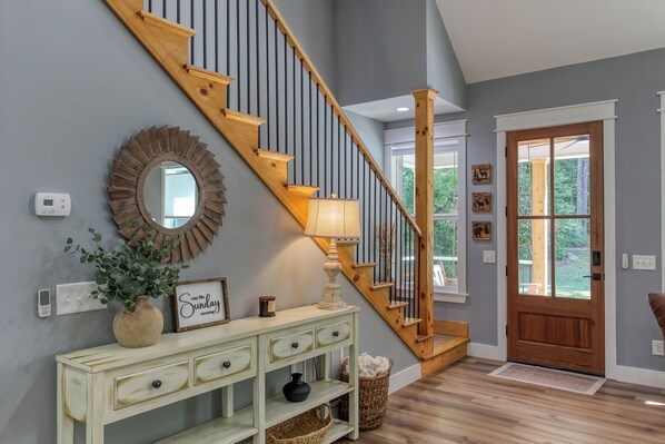 A staircase with wooden steps and black railings, a console table with drawers, a mirror, a wall-mounted thermostat, and a door with a window. - Robbin’s Nest on Snowbird Creek | Fire Pit (Robbinsville)
