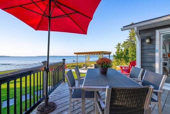 A deck with a table and chairs, a red umbrella, and a view of the water. - Luxury 3BR Lakefront Cottage W/ Sauna (Port McNicoll)