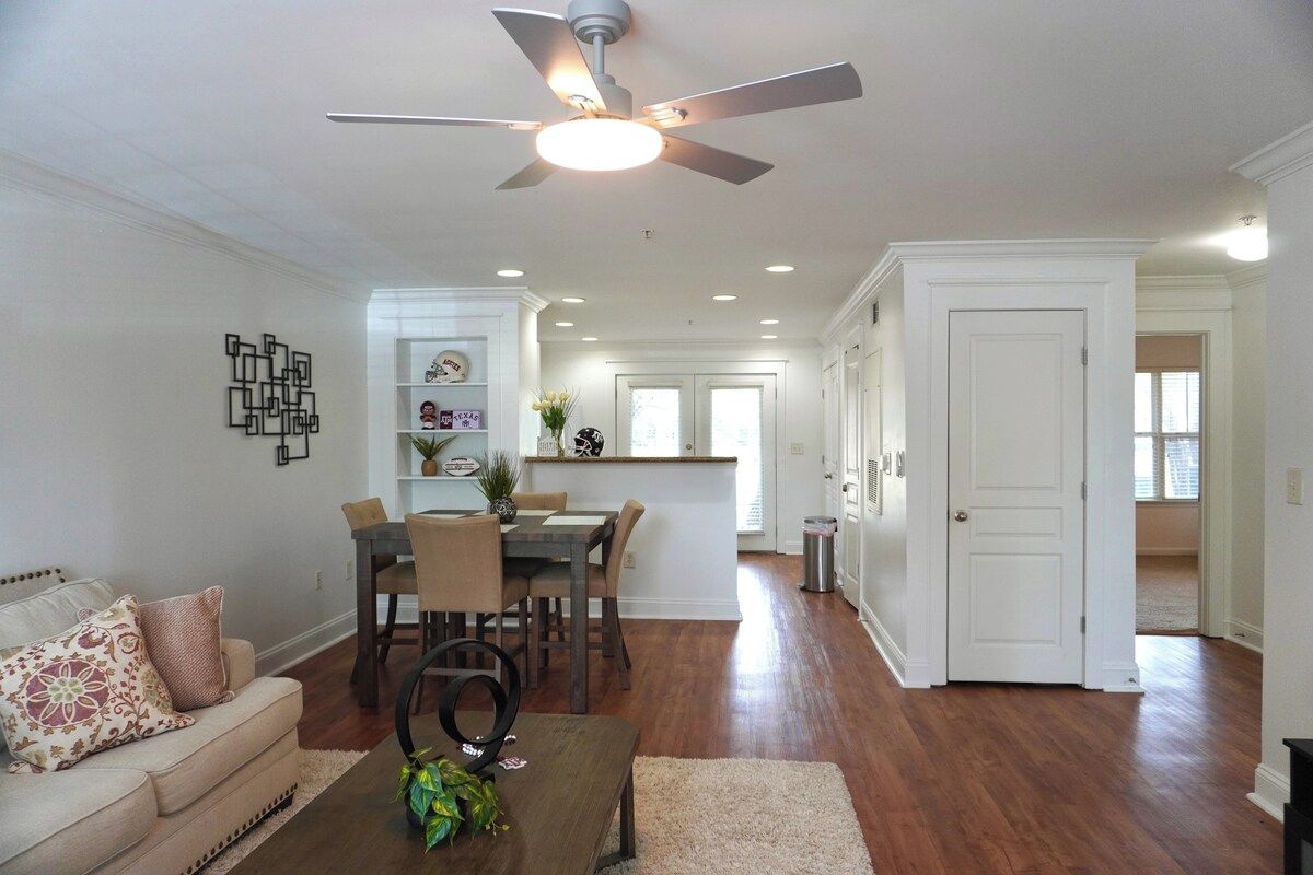 A living room with a ceiling fan, a dining area with chairs and a table, and a kitchen in the background.