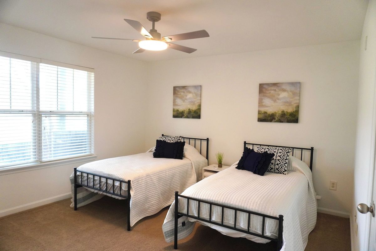 A bedroom with two beds, a ceiling fan, and two wall-mounted paintings.