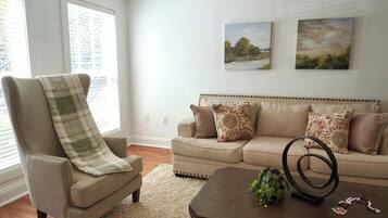 A living room with a beige armchair, a sofa, a coffee table, and two landscape paintings on the wall.