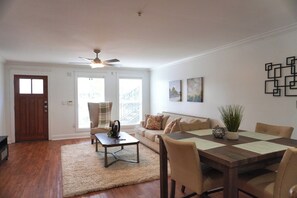 A living room with a wooden floor, a ceiling fan, a sofa, a coffee table, and a dining table with chairs. - Aggieland Getaway  2BR/2BA • Sleeps 6 (College Station)
