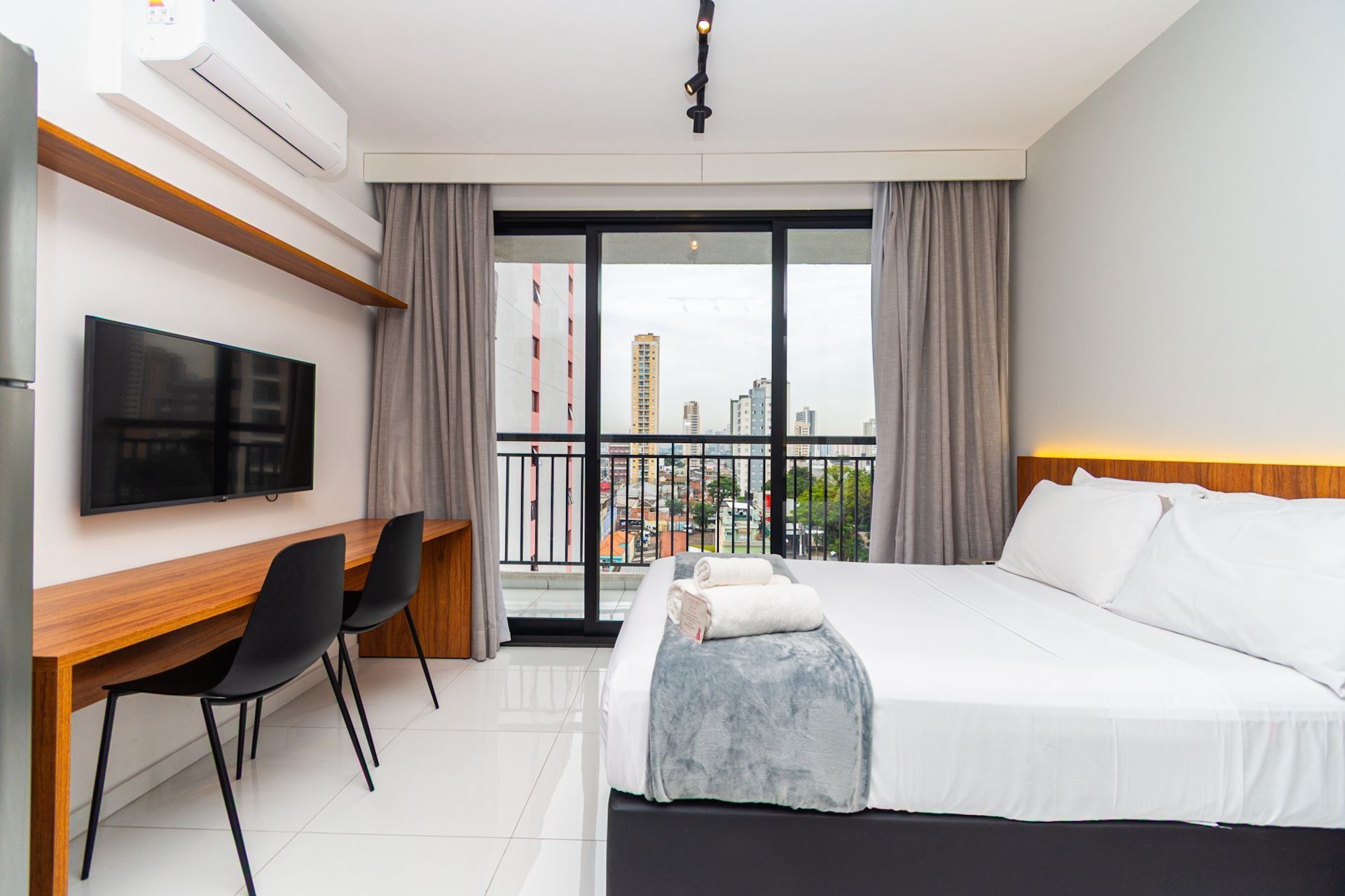 Habitación de hotel con cama, escritorio, silla, televisor y balcón con vista a la ciudad.