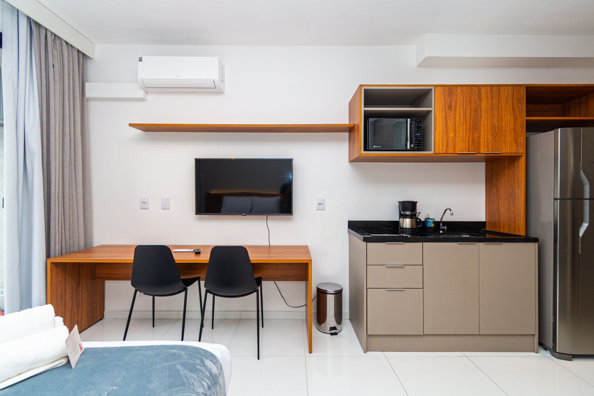Quarto de hotel compacto com cozinha compacta, cama, TV e escrivaninha com duas cadeiras.