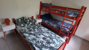 Una cama litera con un edredón estampado con motivos tropicales, una mesita de noche con una lámpara y una pared blanca.