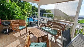 Un patio techado al aire libre con muebles de madera, una piscina y vistas a zonas verdes y colinas.