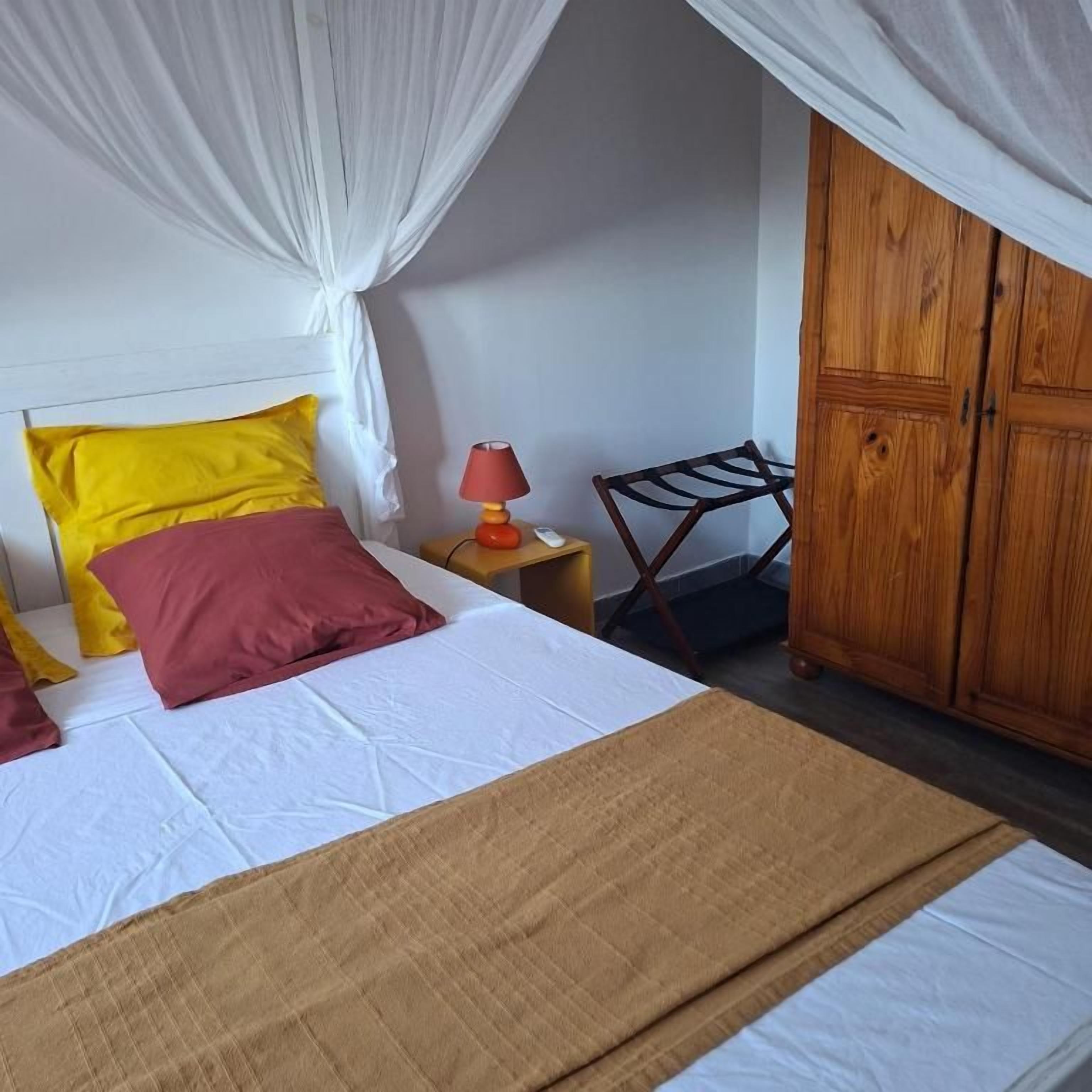 Una cama con colcha blanca y marrón, almohada amarilla y roja, lámpara roja sobre una mesita de noche de madera, y un armario de madera.