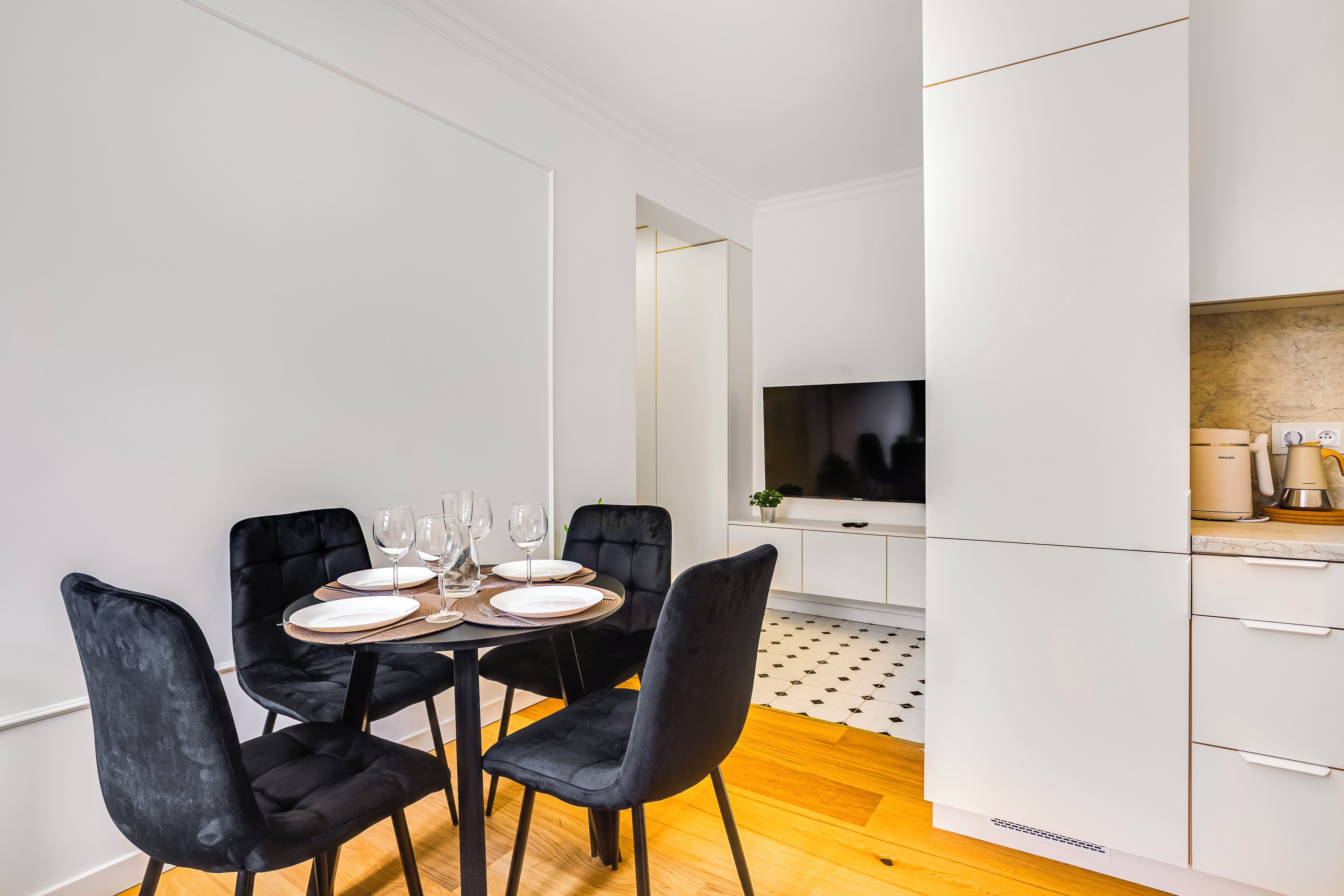 A modern dining area with a table set for four, black chairs, and a kitchen with white cabinets and a large screen.