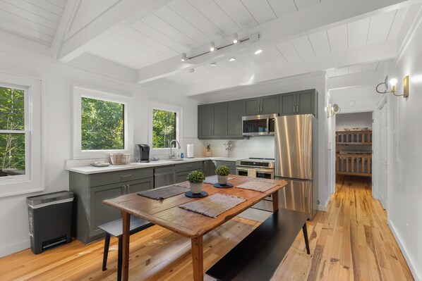 A modern kitchen with a wooden dining table, grey cabinets, stainless steel appliances, and large windows. - Ski Pet Friendly 3BR w/ Deck + Resort Access (Sapphire)