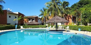 A swimming pool with lounge chairs and thatched umbrellas, surrounded by palm trees and a building with satellite dishes. - CASA MELO 9 - UBATUBA (Ubatuba)