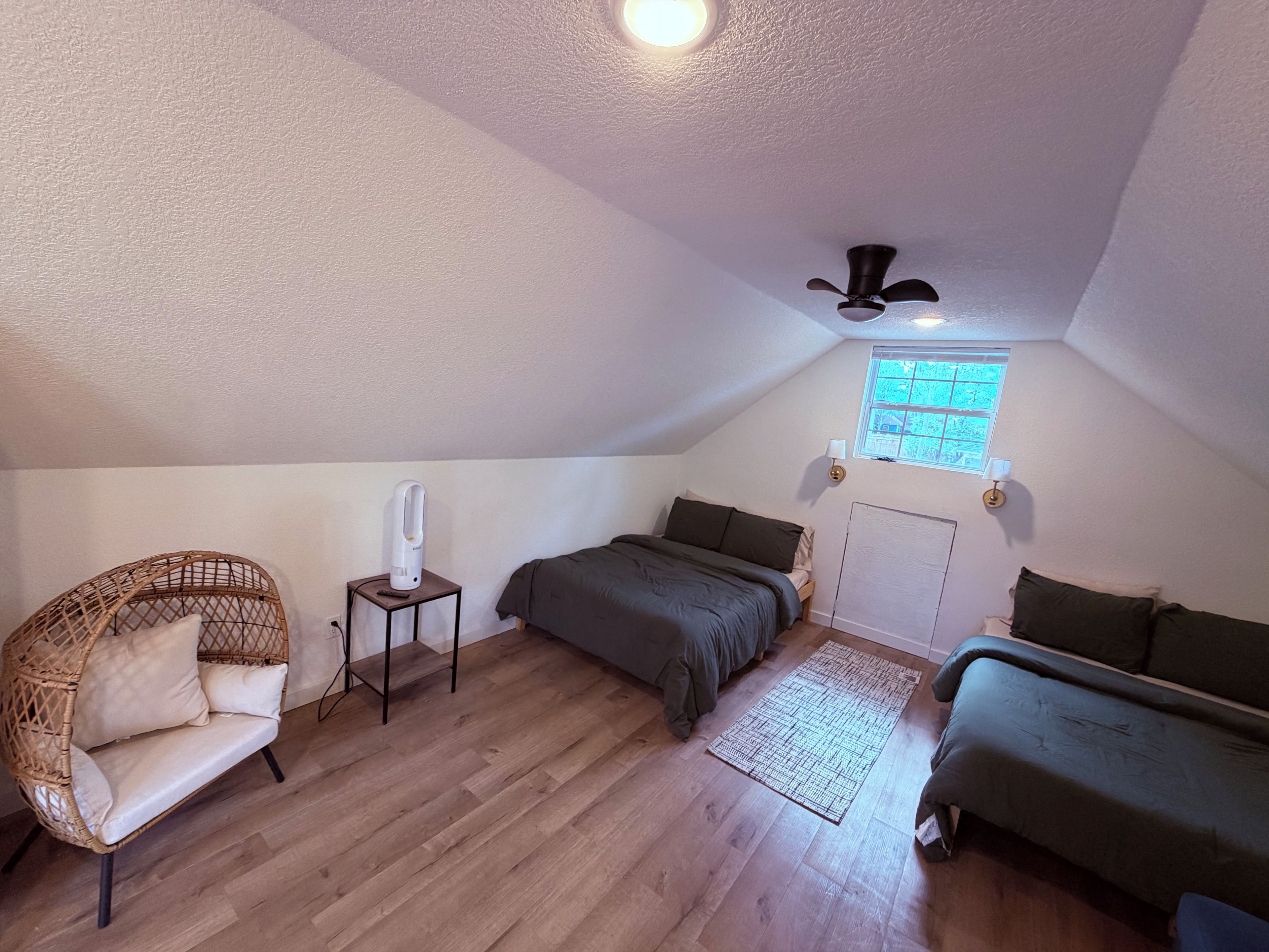 Une chambre mansardée chaleureuse, avec un plafond en pente, un ventilateur de plafond, une fenêtre offrant une belle vue, un lit avec un matelas de couleur sombre, une chaise en osier et une petite table avec une lampe.