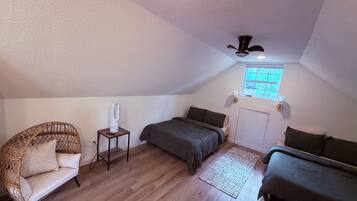 Une chambre mansardée chaleureuse, avec un plafond en pente, un ventilateur de plafond, une fenêtre offrant une belle vue, un lit avec un matelas de couleur sombre, une chaise en osier et une petite table avec une lampe.