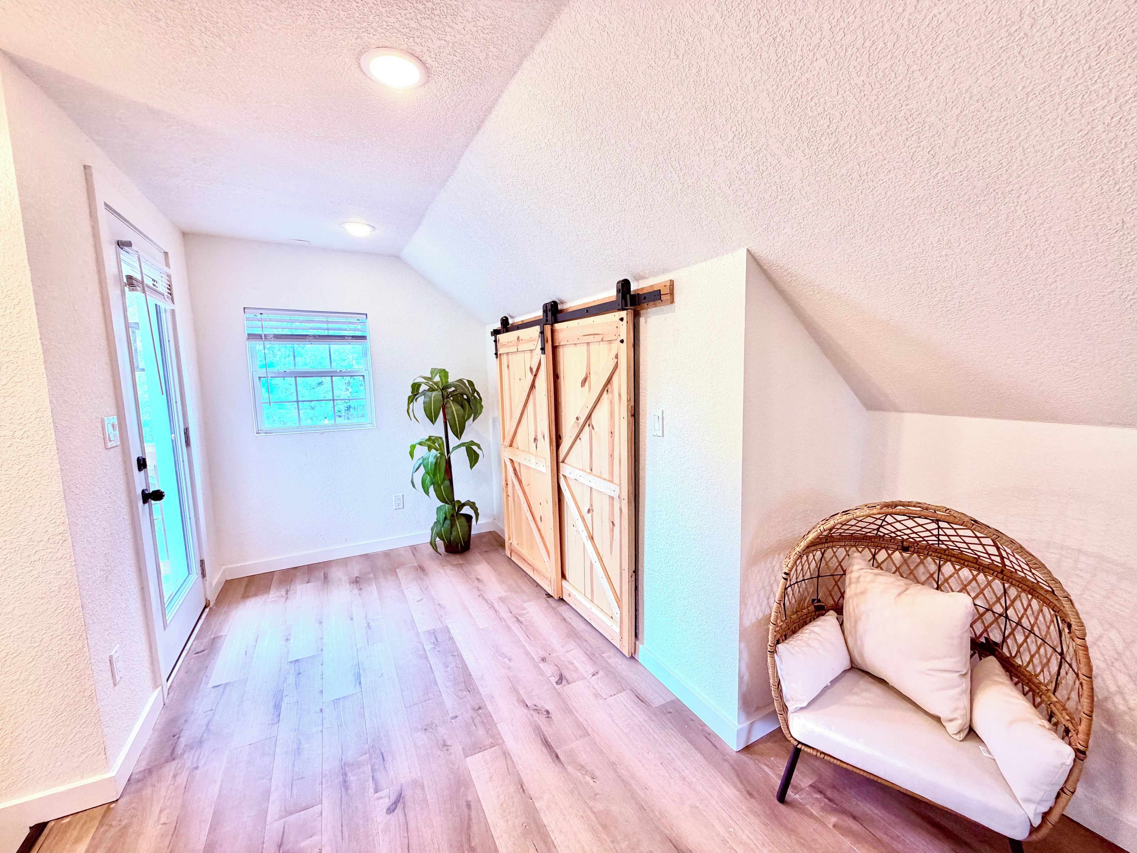 Une pièce avec un plancher en bois, une porte coulissante de style rustique, une plante en pot, un fauteuil en osier avec des coussins et une fenêtre avec des stores.