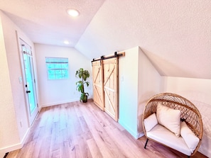 A room with a wooden floor, a sliding barn door, a potted plant, a wicker chair with cushions, and a window with blinds. - Tiny homelWalk to DT Rogers lRailyard Bike Park & Live Musicl EV chargers avail. (Rogers)