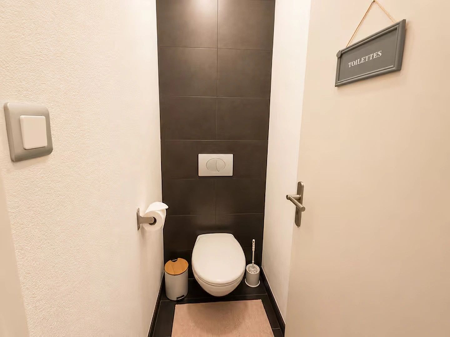 Une petite salle de bain étroite, équipée de toilettes, d’une poubelle et d’une brosse à récurer. Un panneau indique l’emplacement des toilettes.