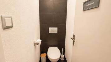 Une petite salle de bain étroite, équipée de toilettes, d’une poubelle et d’une brosse à récurer. Un panneau indique l’emplacement des toilettes.