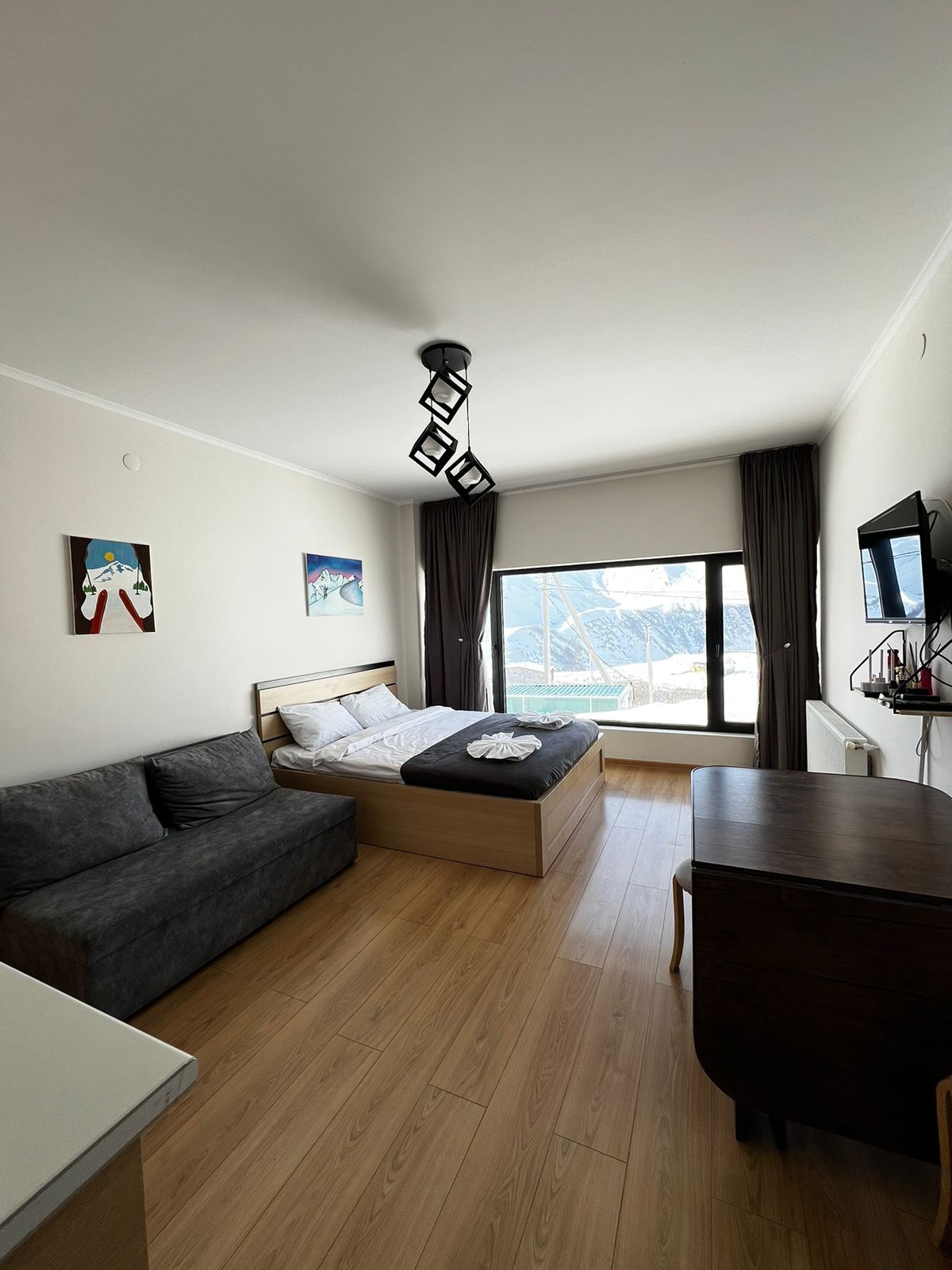 A modern hotel room with a bed, a sofa, a dining table, and a large window offering a view of the outdoors.