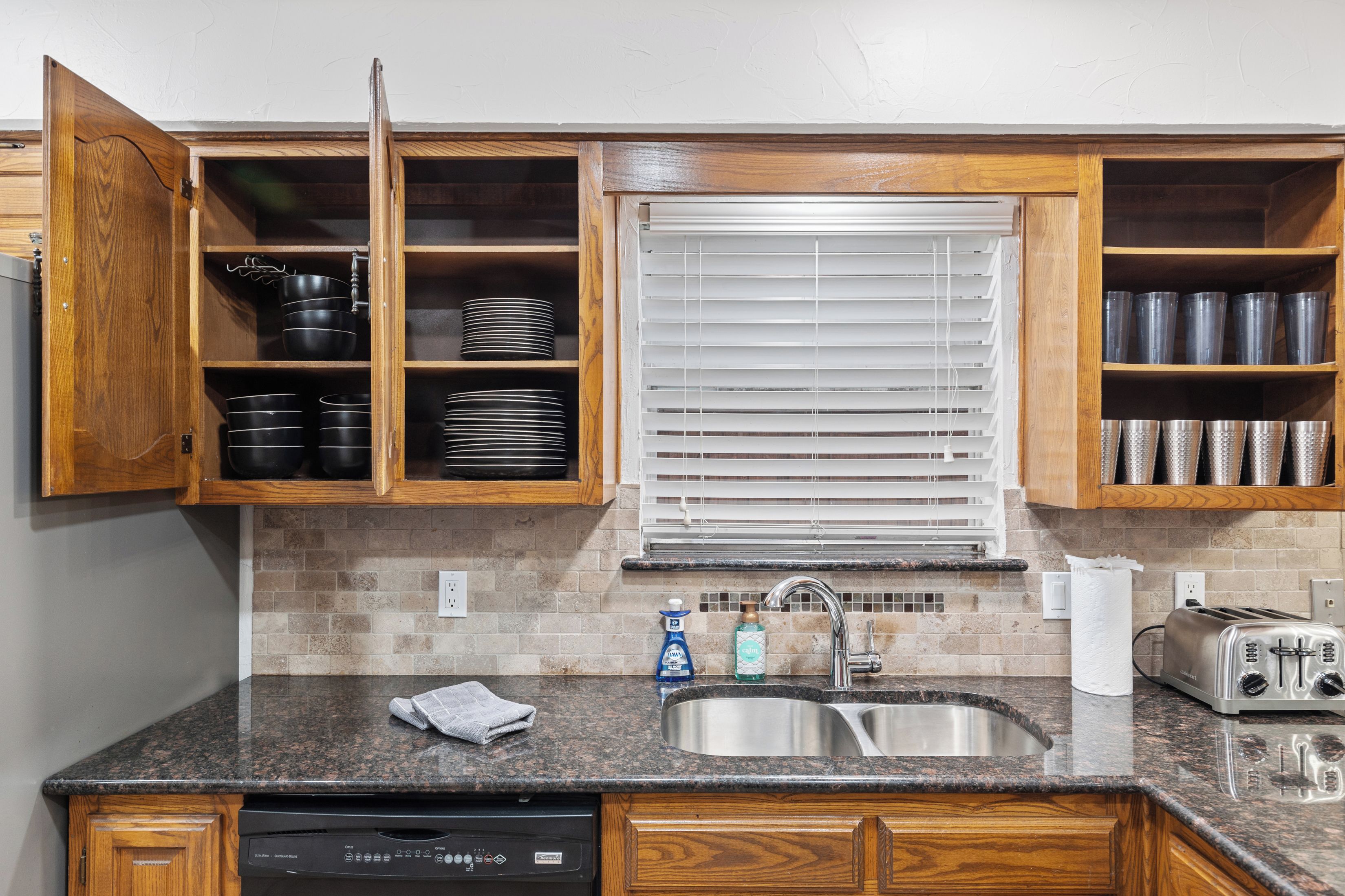 A kitchen with wooden cabinets, a granite countertop, a stainless steel sink, and a built-in window with blinds.