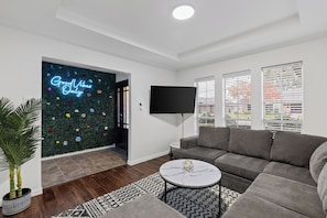 A modern living room with a grey sofa, a round coffee table, a flat-screen TV, and a decorative wall with a 'Good Vibes Only' sign. - Backyard Oasis w/ Pool + Spa and Huge Game Room (Richardson)