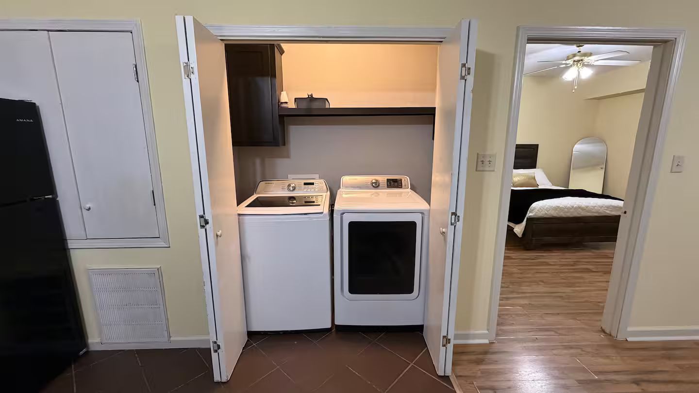 A compact laundry room with two washing machines and a dryer, a small cabinet above, and a bedroom visible through an open door.