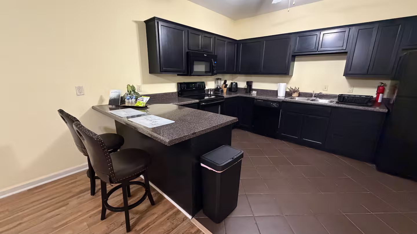 A kitchen with dark cabinets, a granite countertop, a black trash bin, and a bar stool.