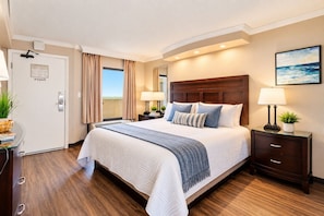 A hotel room with a large bed, a wooden headboard, a nightstand with a lamp, a desk with a computer, and a view of the sea through the window. - Caravelle Resort 1234 - Couples Oceanfront Escape with Pools and Hot tubs (Myrtle Beach)