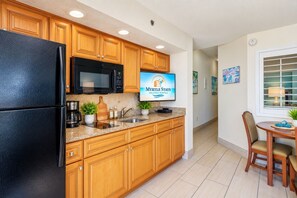 Cozinha com armários de madeira, geladeira preta, micro-ondas e uma televisão exibindo 'Myrtle Stays Vacation Rentals'.