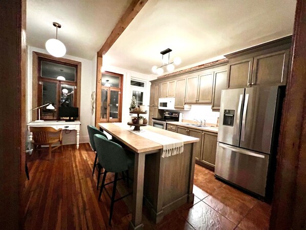 A kitchen with wooden cabinets, a stainless steel refrigerator, a microwave, and a wooden dining table with chairs. - Urban Living at Cartier – A Condo in the Heart of the Arts (Québec)