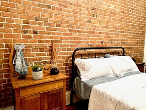 A bedroom with a brick wall, a wooden nightstand, a bed with white bedding, a lamp, and a polka-dotted towel. - Urban Living at Cartier – A Condo in the Heart of the Arts (Québec)