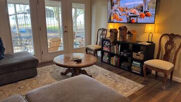 A living room with a television, ottomans, a bookshelf, and a view through the glass doors.