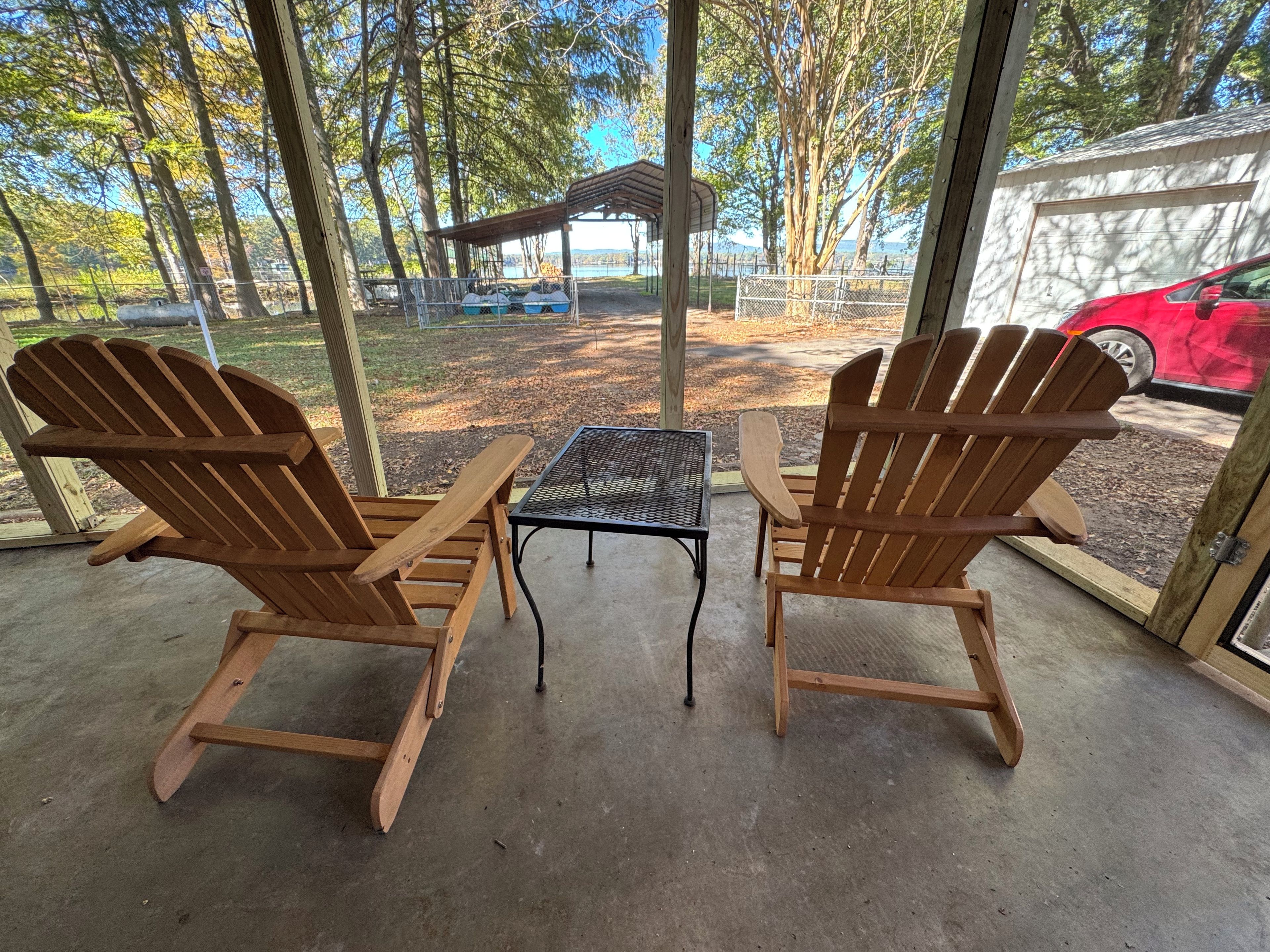 Two wooden Adirondack chairs facing a metal table and a view of a park with a covered area and a red car parked outside.