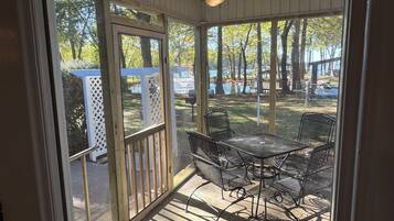 A covered patio with metal chairs and a table, overlooking a wooded area with a lake.
