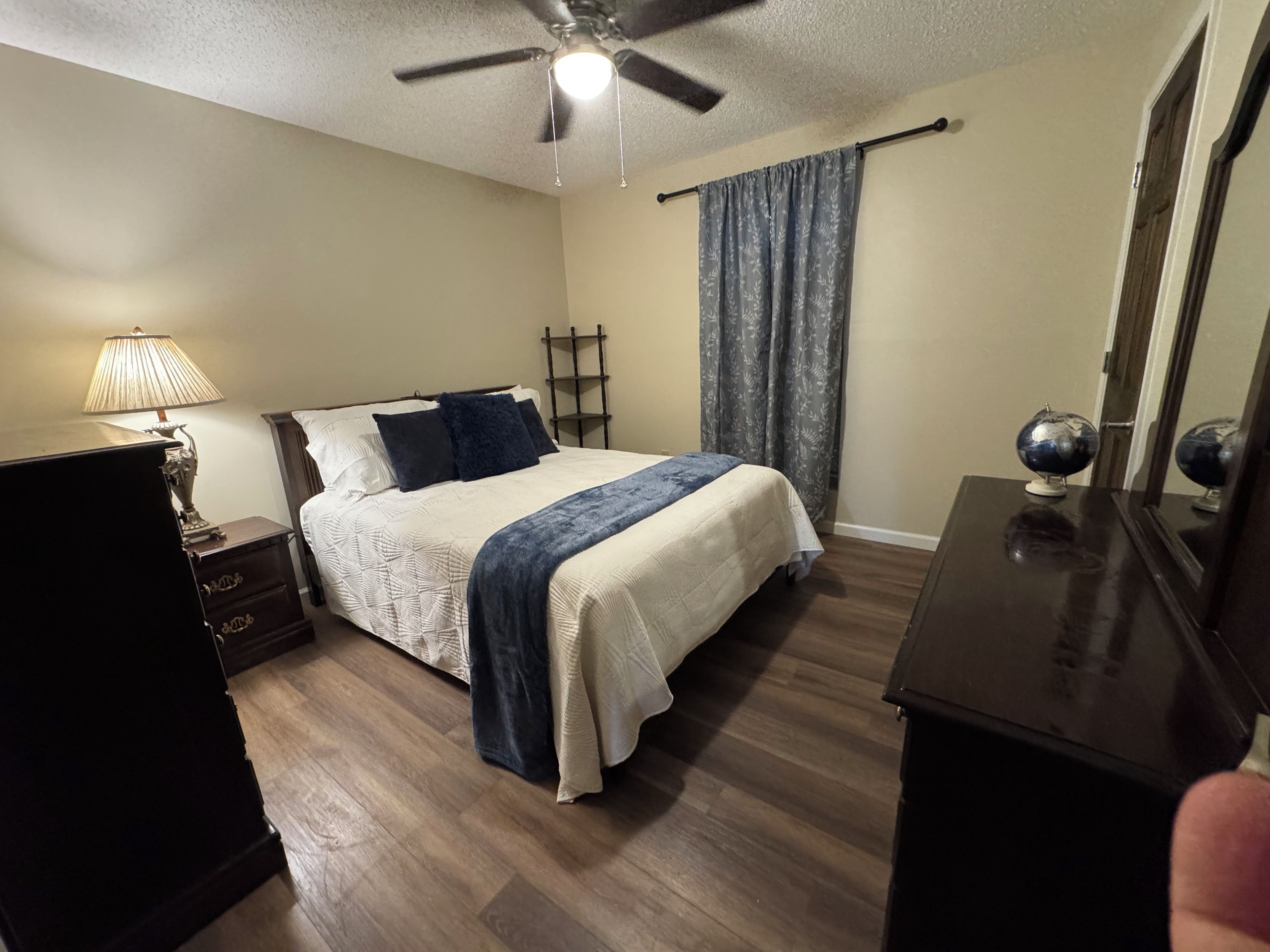 A bedroom with a bed, a nightstand, a lamp, a ceiling fan, a window with curtains, and a dresser.