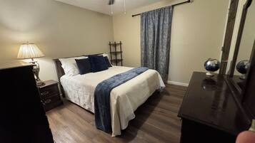 A bedroom with a bed, a nightstand, a lamp, a ceiling fan, a window with curtains, and a dresser.