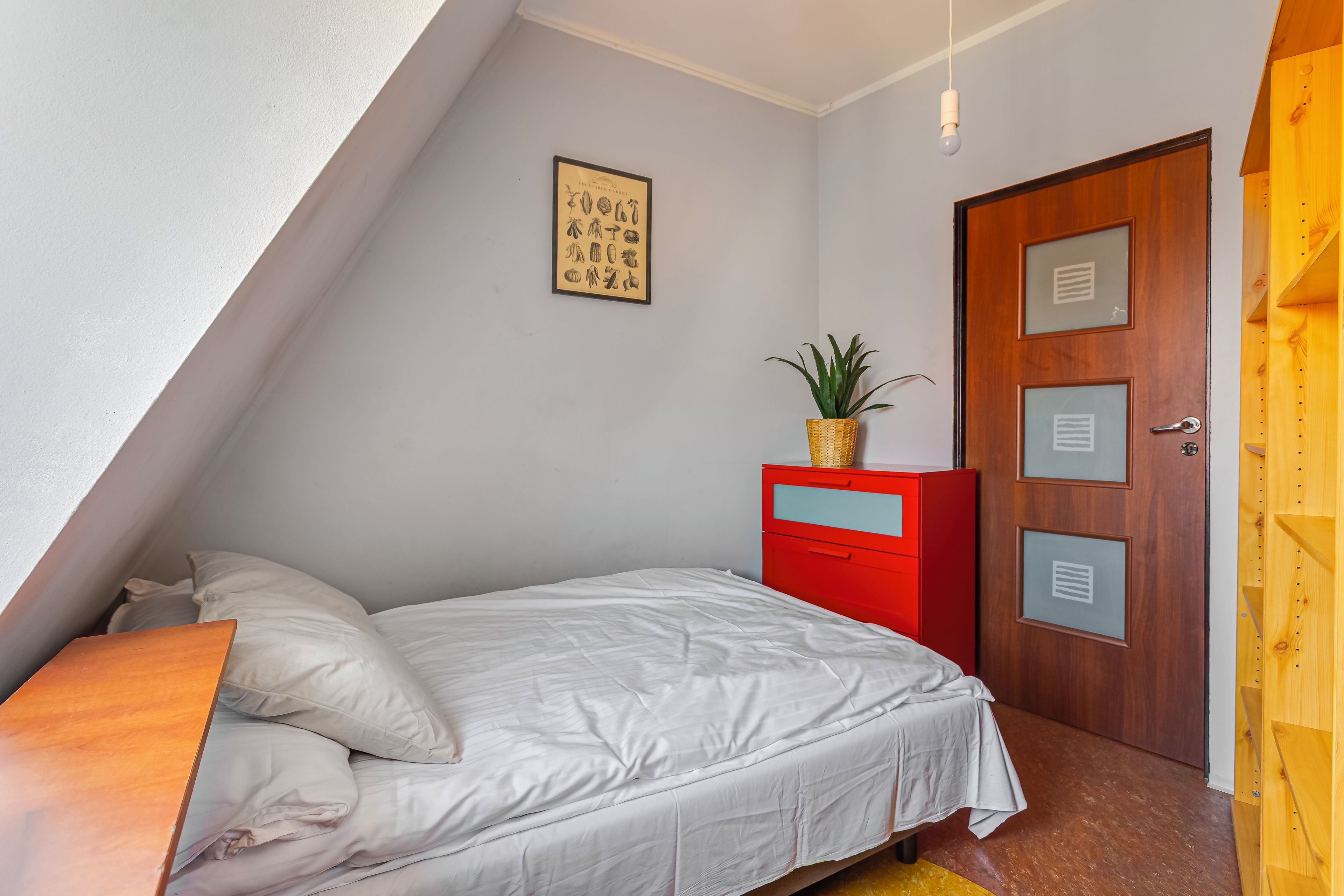 Un dormitorio con una cama, un aparador rojo, una puerta de madera e ilustración botánica enmarcada en la pared.