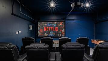 A dark room with a large screen displaying 'NETFLIX' and a selection of movies. There are black leather chairs arranged in front of the screen.