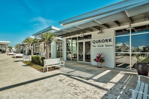 A storefront with a sign reading 'QUEORK The Cork Store' and a paved walkway with benches. - Recovery Room (Santa Rosa Beach)