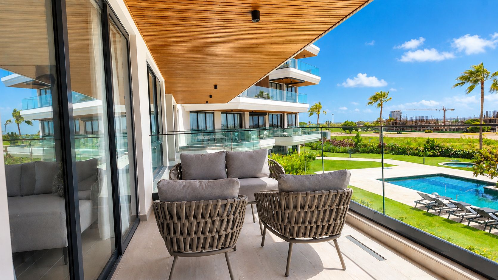 A balcony with wicker furniture, a glass balustrade, and a view of a swimming pool and greenery.