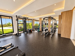 A modern gym with various exercise equipment, large windows, and a view of greenery outside.