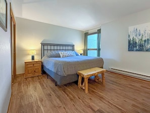 Quarto com cama, mesas de cabeceira, um banco, janela com cortinas e um quadro na parede.