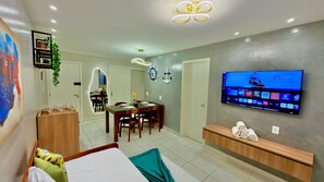 Habitación de hotel moderna con zona de comedor, un televisor mostrando un canal y un estante en la pared con toallas.