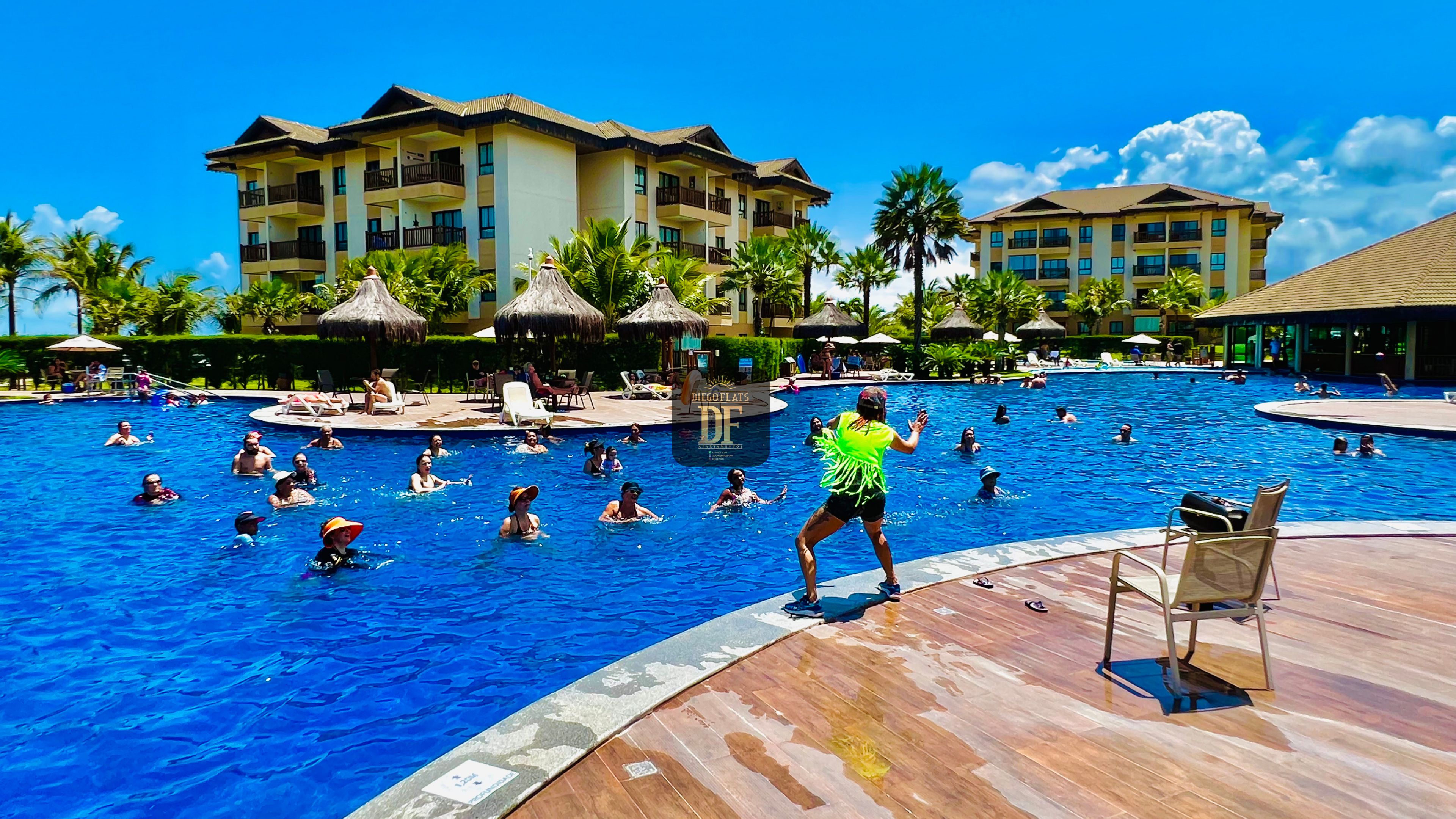 Una piscina grande con gente nadando y tomando el sol, rodeada de palmeras y un edificio de resort.