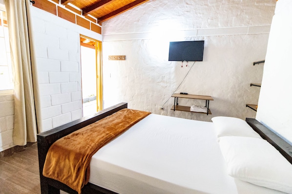 Une pièce comprenant un lit, un téléviseur fixé au mur, une étagère en bois et une fenêtre avec des rideaux.