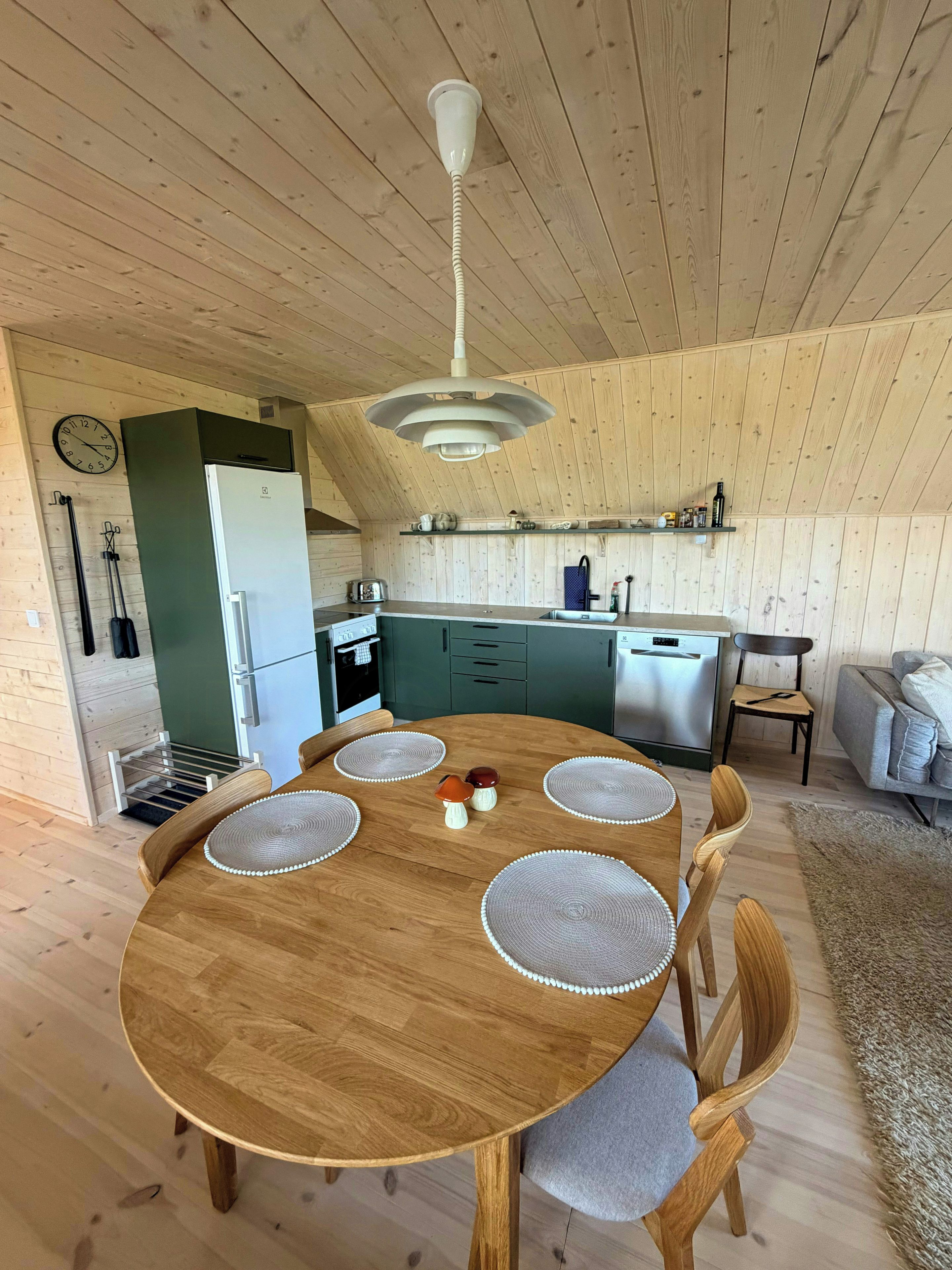A wooden dining table with placemats and chairs, a kitchen with a built-in oven and sink, and a hanging light fixture.