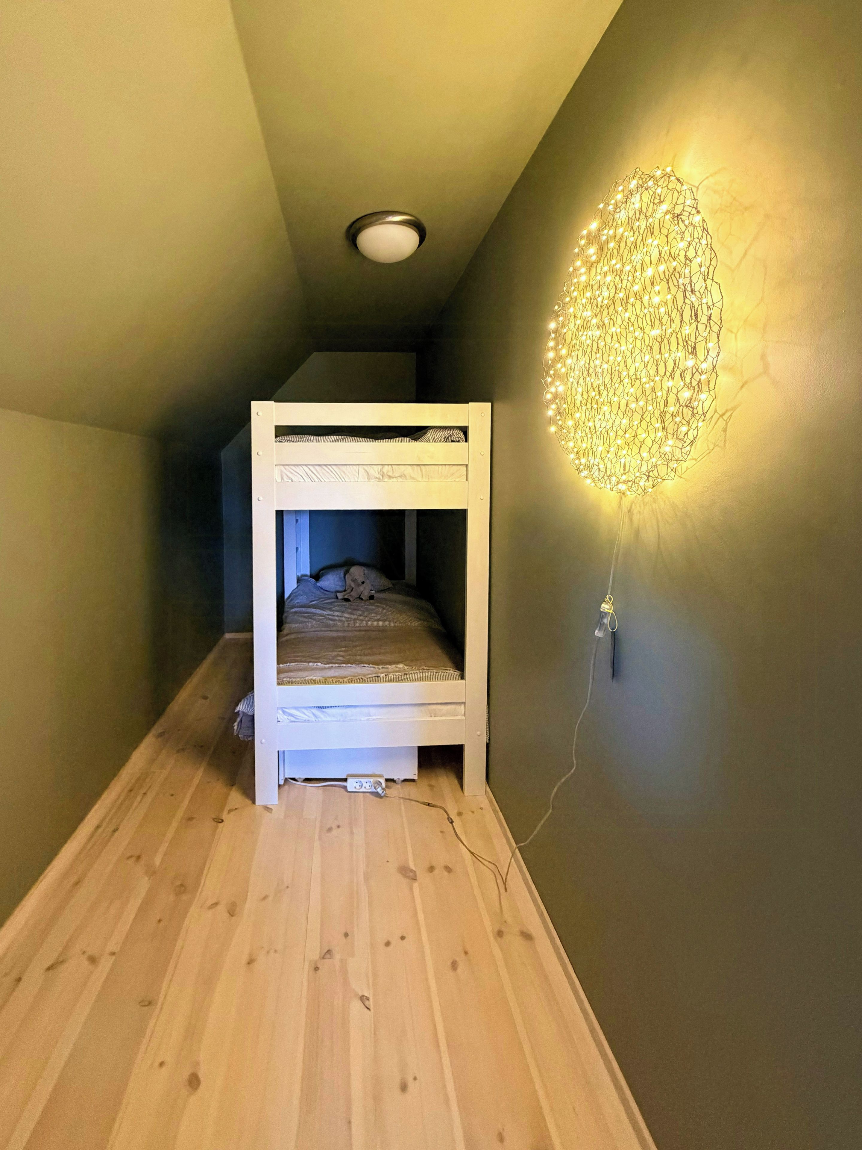 Un lit superposé dans une pièce étroite, avec un plancher en bois, un luminaire fixé au mur et un plafonnier.
