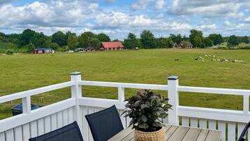 A wooden deck with a table and chairs, a potted plant, and a view of a grassy field with sheep and red buildings in the distance.